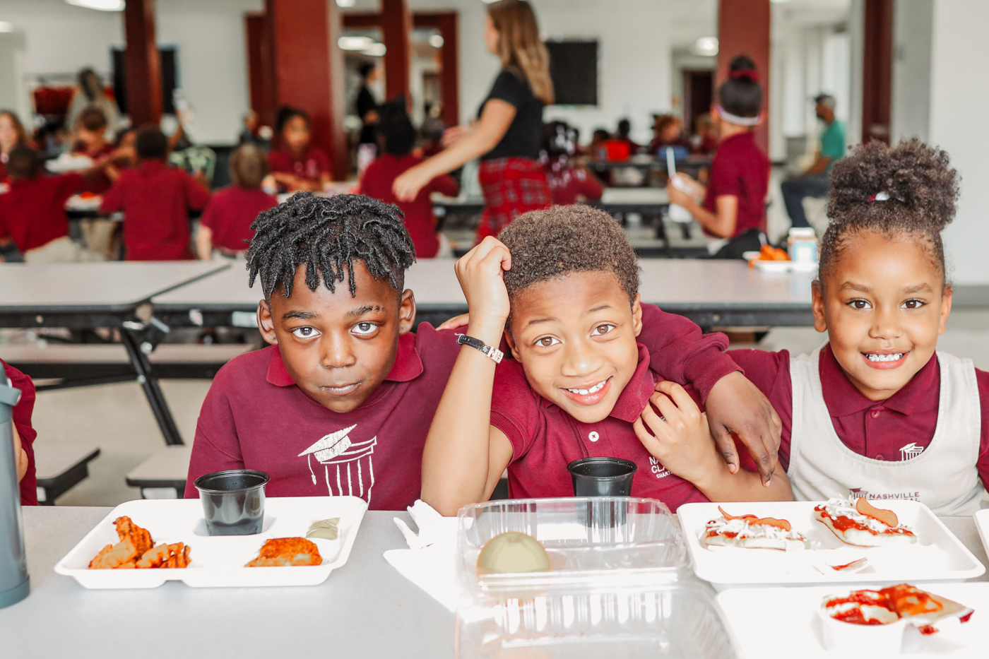3 kids sit together smiling and enjoying lunch in the cafeteria