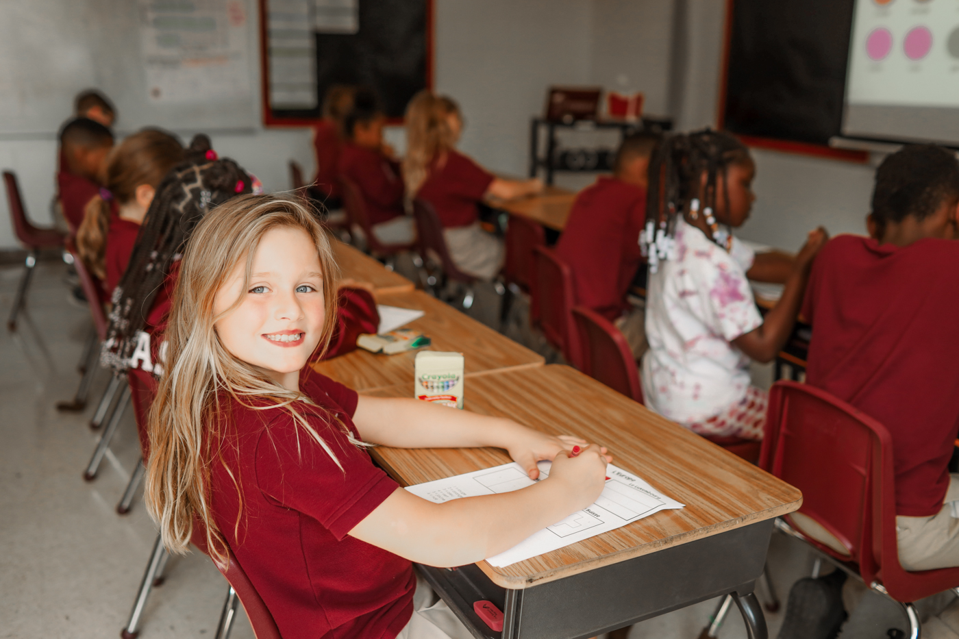 Young girl sits at desk with crayon and paper smiling at camera
