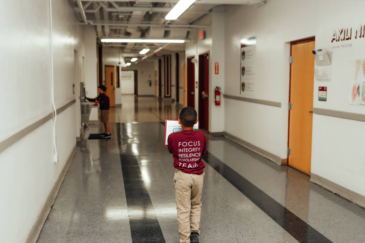 Child is walking through school hallway holding book, with his back to the camera the shirt reads "focus, integrity, resilience, scholarship, team"