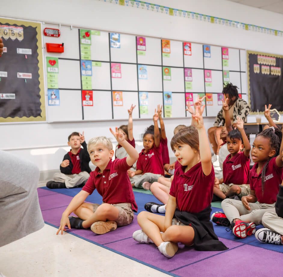 Young students sit on brightly colored flooring raising hands