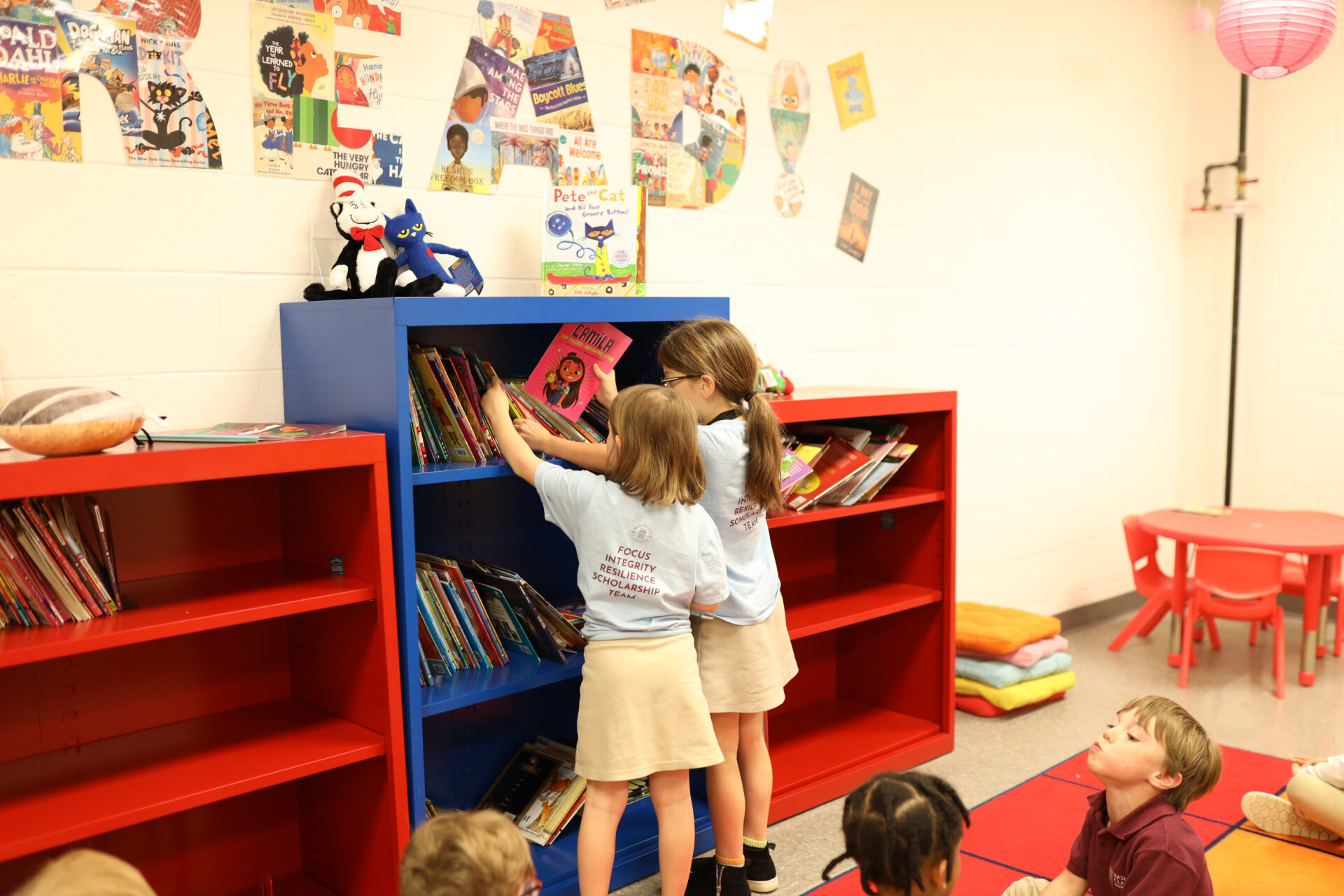 Two students with backs facing the camera put books up in primary colored book shelves.